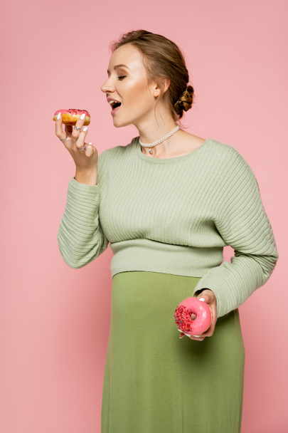 Terhes nő zöld pulóverben, rózsaszín fánkot tart.  - Fotó, kép