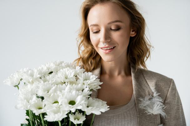 Stylish woman in jacket looking at bouquet of white flowers isolated on grey  - Photo, Image