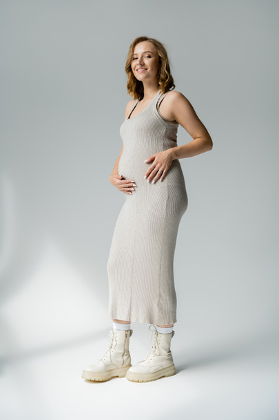 Volle Länge der stilvollen schwangeren Frau im Kleid, die in die Kamera auf grauem Hintergrund schaut  - Foto, Bild
