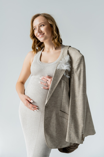 Модная беременная женщина в платье и пиджаке, касающаяся живота, изолированного на серой  - Фото, изображение