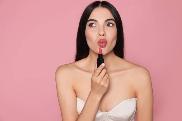 Emotional young woman with beautiful makeup holding glossy lipstick on pink background - Foto, Imagem