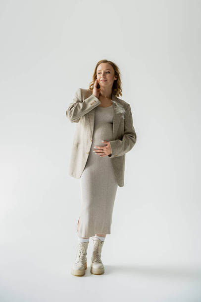 Pleine longueur de femme enceinte à la mode en bottes et veste posant sur fond gris  - Photo, image