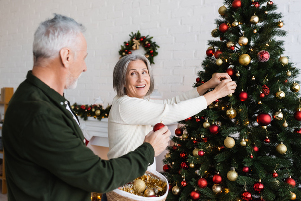 χαρούμενη μεσήλικη γυναίκα με γκρίζα μαλλιά διακοσμώντας χριστουγεννιάτικο δέντρο και κοιτάζοντας το σύζυγο κρατώντας ψάθινο καλάθι  - Φωτογραφία, εικόνα