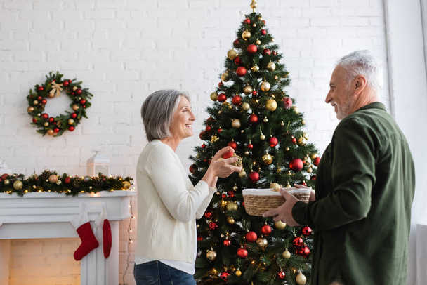 smiling mature woman with grey hair decorating christmas tree and looking at husband holding wicker basket  - Fotografie, Obrázek