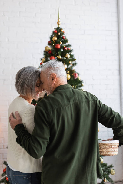 χαμογελαστό ζευγάρι μέσης ηλικίας αγκαλιάζει ενώ διακόσμηση χριστουγεννιάτικο δέντρο - Φωτογραφία, εικόνα