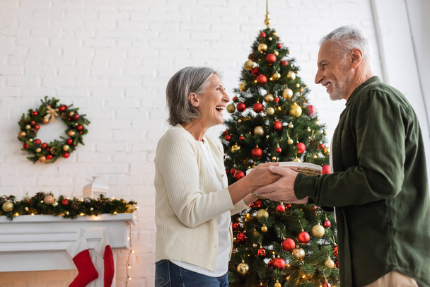 glückliches Paar mittleren Alters hält Weidenkorb neben geschmücktem Weihnachtsbaum - Foto, Bild