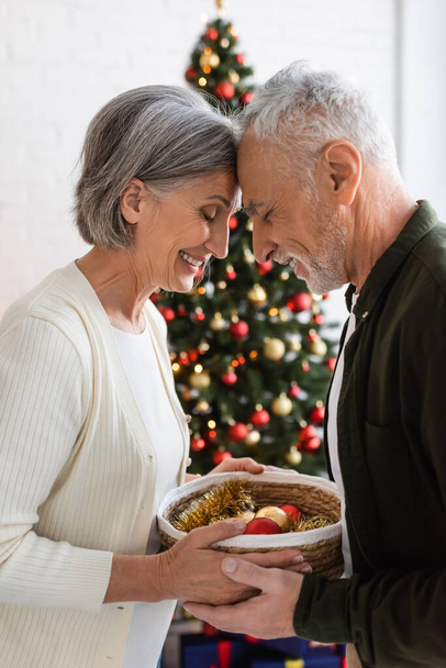 sonriente pareja de mediana edad con los ojos cerrados sosteniendo canasta de mimbre cerca de árbol de Navidad decorado - Foto, imagen