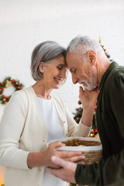 happy mature couple with closed eyes holding wicker basket near decorated christmas tree - Photo, Image