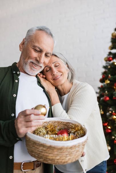 Lächelnde Frau mittleren Alters lehnt mit Weidenkorb am Weihnachtsbaum an Ehemann - Foto, Bild