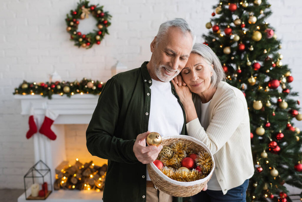 radosna kobieta w średnim wieku opierając się na mężu z wiklinowym koszem patrząc na bal bożonarodzeniowy - Zdjęcie, obraz