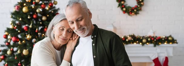 cheerful middle aged woman leaning on husband near decorated christmas tree, banner - Photo, Image