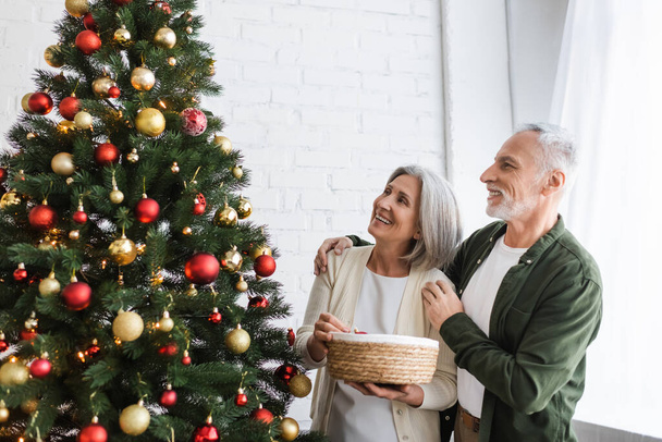 alegre mediana edad marido y esposa mirando decorado árbol de Navidad - Foto, imagen
