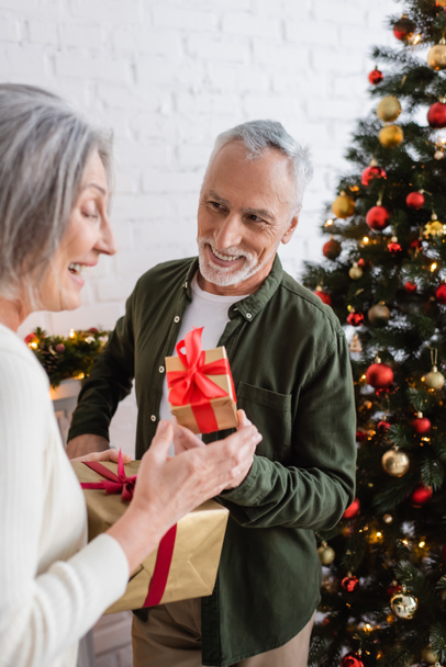 szczęśliwy mężczyzna w średnim wieku trzyma pudełko prezentów i patrząc na żonę w pobliżu choinki - Zdjęcie, obraz