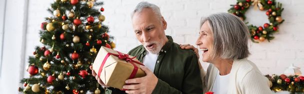 mujer madura feliz mirando sorprendido marido con regalo de Navidad cerca de abeto decorado, pancarta - Foto, imagen