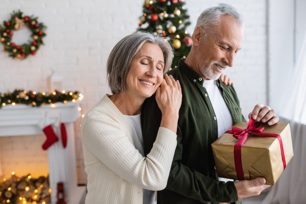 uśmiechnięta kobieta z zamkniętymi oczami przytulająca dojrzałego brodatego męża z prezentem świątecznym  - Zdjęcie, obraz