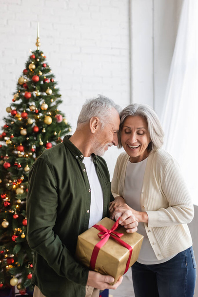 bearded middle aged man holding present and hugging happy wife near christmas tree - Foto, Imagem