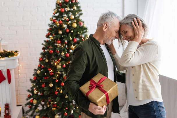 bärtiger Mann mittleren Alters hält Geschenk in der Hand und umarmt lächelnde Frau am Weihnachtsbaum - Foto, Bild