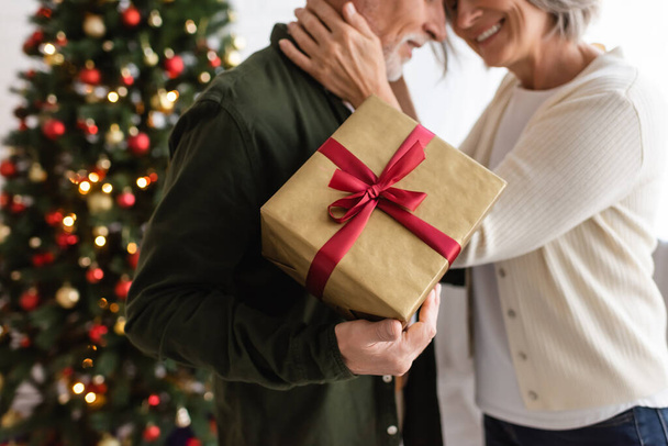 καλλιεργημένη άποψη του γενειοφόρου μεσήλικας άνδρας που κρατά το παρόν και αγκαλιάζει με ευτυχισμένη σύζυγο κοντά στο χριστουγεννιάτικο δέντρο - Φωτογραφία, εικόνα