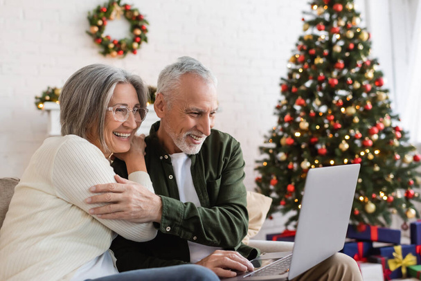 smiling middle aged couple watching comedy movie on laptop near decorated christmas tree  - Фото, зображення