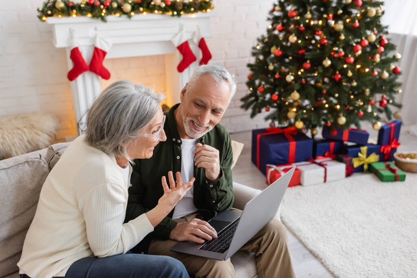 lächelnde Frau mittleren Alters zeigt auf glücklichen Ehemann und hat Videoanruf auf Laptop während Weihnachten  - Foto, Bild