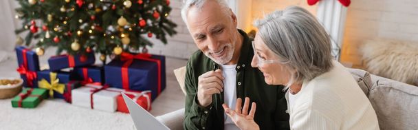 улыбающаяся женщина средних лет, указывающая на счастливого мужа и имеющая видеозвонок на ноутбуке во время Рождества, баннер - Фото, изображение