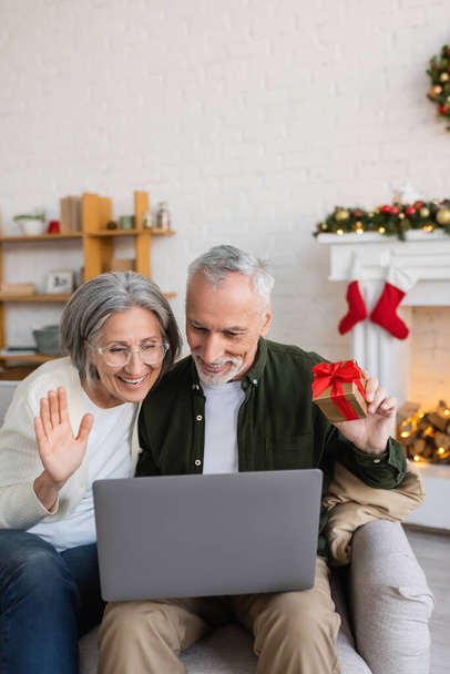 smiling middle aged woman waving hand near husband and having video call on laptop during christmas  - Photo, Image