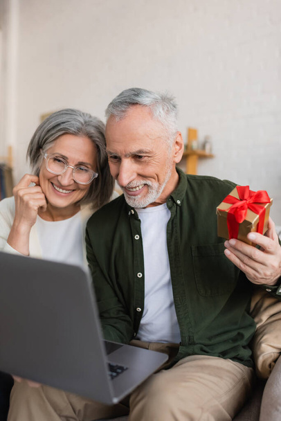 cheerful middle aged man holding christmas present near wife during video call - Foto, Bild