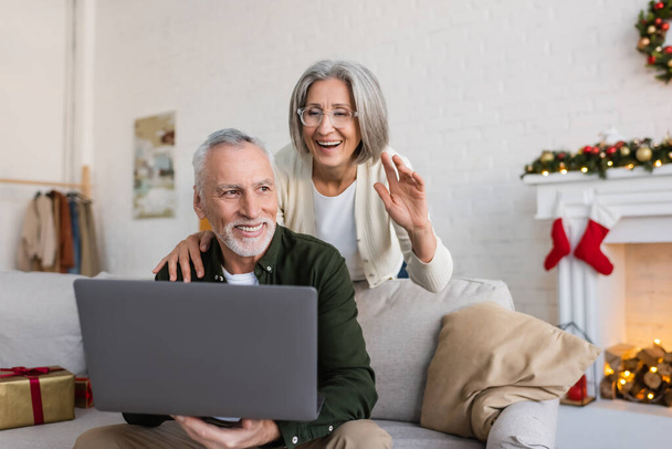 fröhliche Frau mittleren Alters in Brille winkt ihrem Mann während eines Videotelefonats am Weihnachtstag die Hand - Foto, Bild