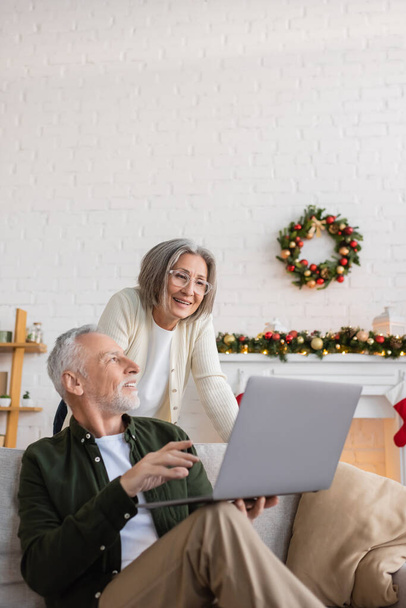 alegre hombre de mediana edad mostrando portátil a la esposa en gafas en el día de Navidad  - Foto, Imagen