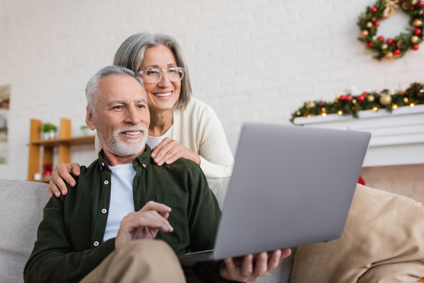 χαρούμενη μεσήλικη γυναίκα με γυαλιά αγκαλιάζει τον σύζυγο και κοιτάζει το λάπτοπ κατά τη διάρκεια βιντεοκλήσης την ημέρα των Χριστουγέννων  - Φωτογραφία, εικόνα