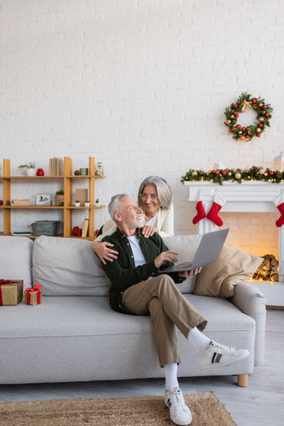szczęśliwy w średnim wieku kobieta w okularach przytulanie mąż siedzi na kanapie z laptopem w Boże Narodzenie  - Zdjęcie, obraz