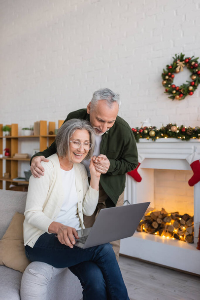 веселая женщина средних лет в очках с помощью ноутбука и держась за руки со счастливым мужем во время рождественских праздников  - Фото, изображение