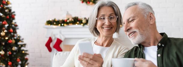 glimlachende en volwassen vrouw in bril houden smartphone in de buurt van echtgenoot met kopje thee, banner - Foto, afbeelding