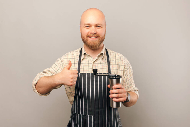 Joven camarero calvo sostiene y recomienda un molinillo de café con un pulgar hacia arriba sobre fondo gris claro. - Foto, Imagen