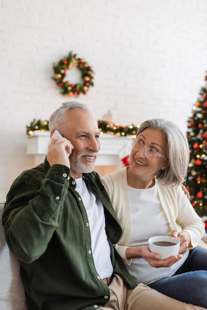 heureux homme d'âge moyen parlant sur smartphone près de la femme avec une tasse de thé pendant Noël  - Photo, image
