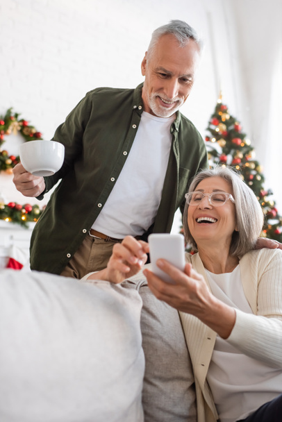 szczęśliwy Dojrzały mężczyzna trzyma filiżankę herbaty i patrząc na smartfona w pobliżu żony i niewyraźne choinki - Zdjęcie, obraz