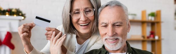 Kredi kartıyla gülümseyen olgun bir kadın sakallı kocasının yanında gülümsüyor. - Fotoğraf, Görsel