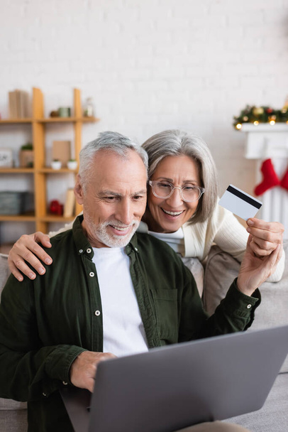 fröhlicher Mann mittleren Alters benutzte zu Weihnachten Laptop in der Nähe einer fröhlichen Frau mit Kreditkarte   - Foto, Bild