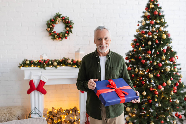 happy middle aged man holding wrapped present near decorated christmas tree  - Valokuva, kuva