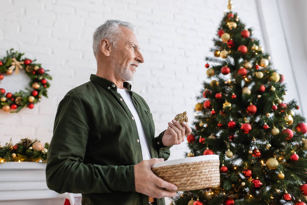 Lächelnder Mann mittleren Alters mit Bart hält Weidenkorb und Christbaumkugeln in der Nähe des Weihnachtsbaums  - Foto, Bild