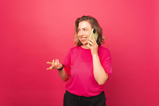 Sourire jeune femme blonde est geste et avoir une conversation sur son téléphone sur fond rose. - Photo, image