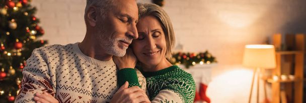 cheerful woman in festive sweater hugging husband near decorated christmas tree in evening, banner - Foto, Bild