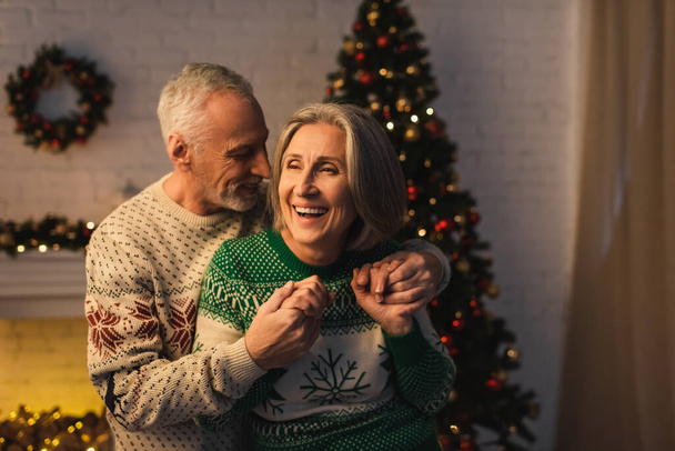 joyeux barbu homme en chandail festif étreignant heureux femme mûre près de l'arbre de Noël dans la soirée - Photo, image