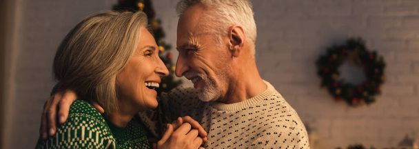 fröhlicher reifer Mann im festlichen Pullover umarmt freudige Frau am Heiligabend, Banner - Foto, Bild
