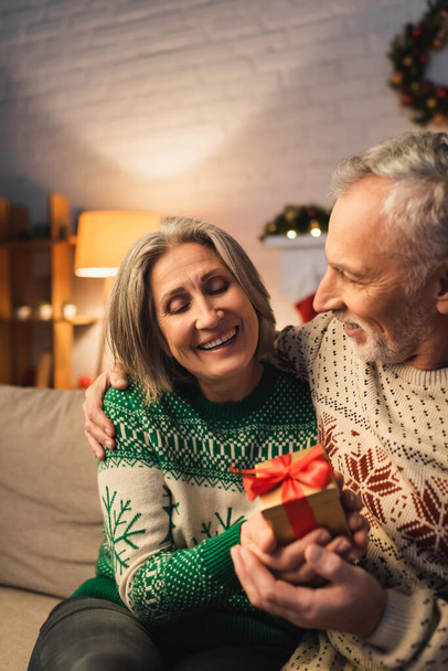 χαρούμενη μεσήλικη γυναίκα που κρατάει χριστουγεννιάτικο δώρο κοντά στον άντρα της με πουλόβερ  - Φωτογραφία, εικόνα