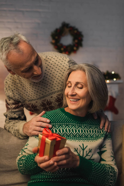 felice donna di mezza età che tiene regalo di Natale vicino al marito in maglione festivo  - Foto, immagini