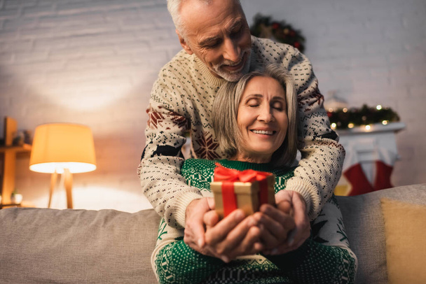 pleased middle aged couple in festive sweaters holding christmas present in hands - Photo, Image
