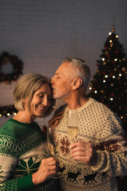 Mann mittleren Alters im festlichen Pullover hält ein Glas Champagner in der Hand und küsst seine glückliche Frau am Weihnachtsbaum - Foto, Bild