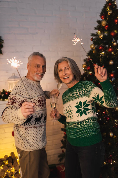 joyful middle aged couple in festive sweaters holding sparklers and glasses of champagne on christmas eve - Photo, Image
