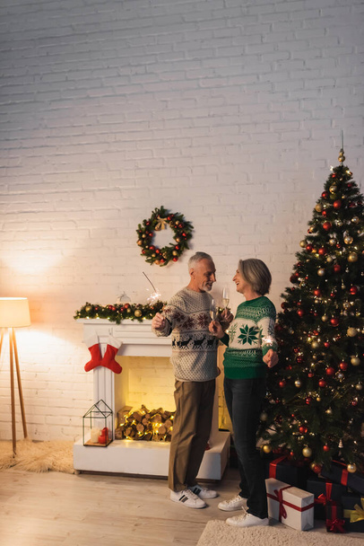 happy mature couple in sweaters holding glasses of champagne and sparklers on christmas eve at home - Photo, Image